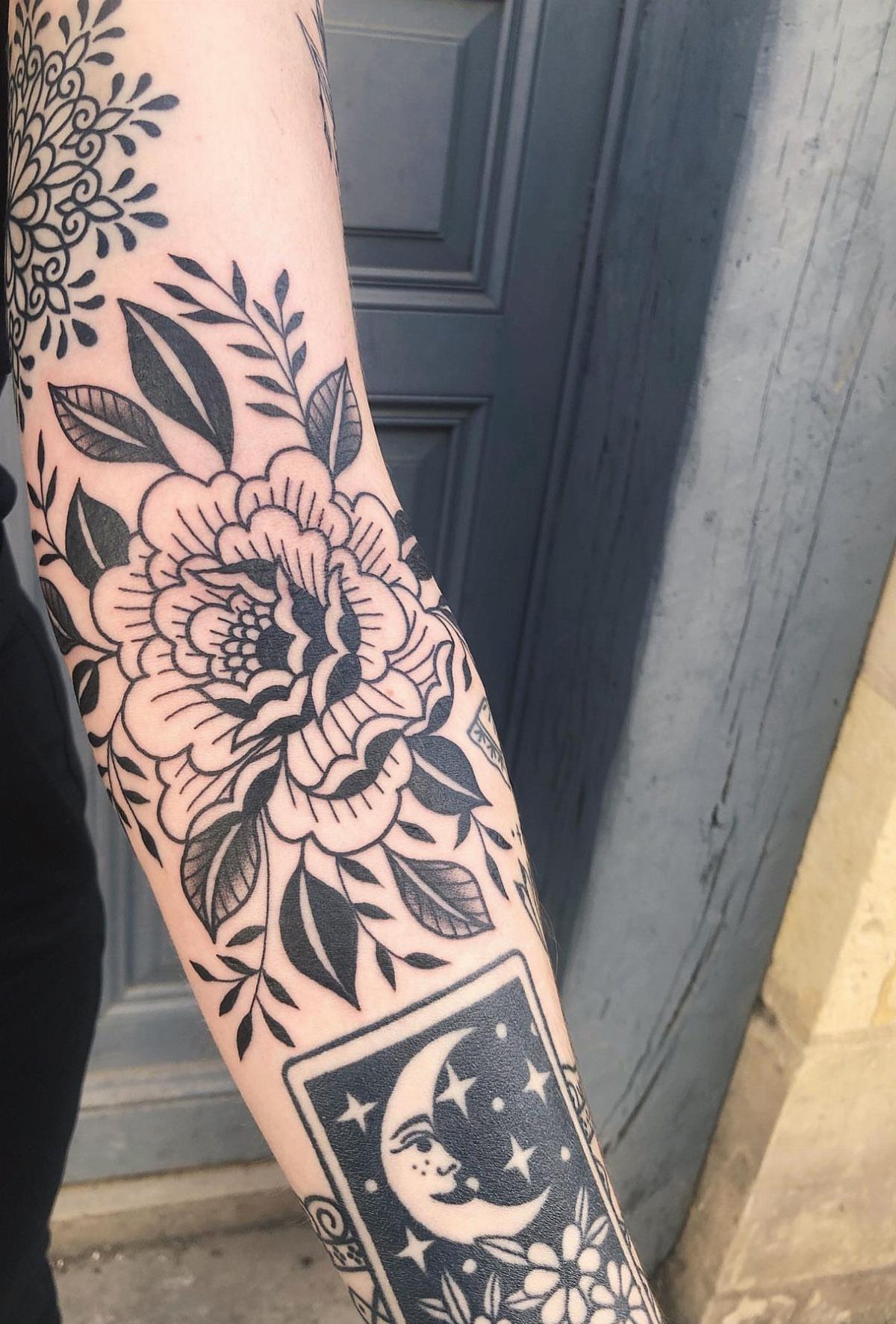 A person's arm decorated with intricate black ink floral and celestial tattoos.