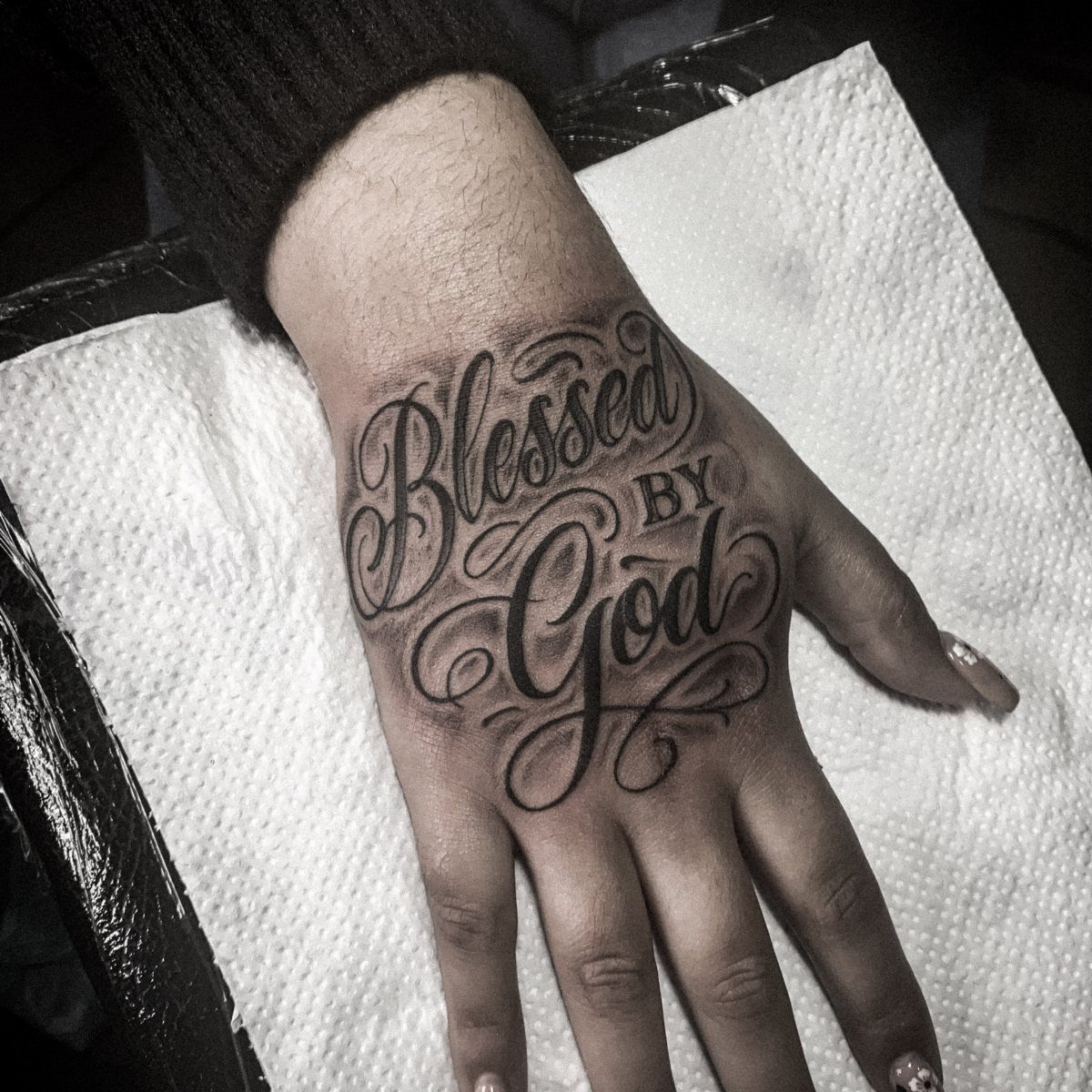 A hand with a large cursive tattoo reading Blessed BY God on the back.