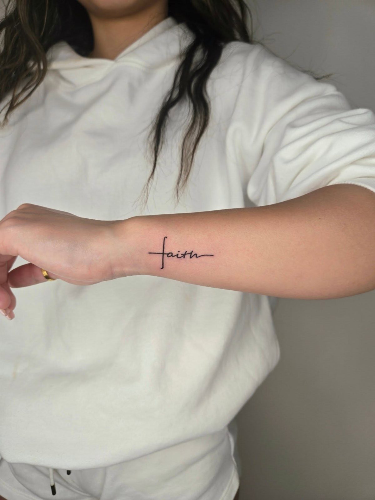 A minimalist black ink tattoo of the word faith integrated with a small cross on a wrist.