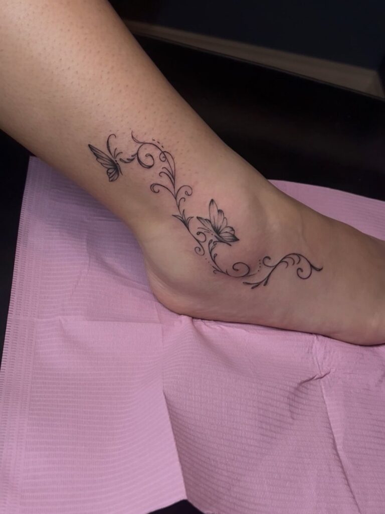 A delicate black ink butterfly and swirl tattoo on a person's ankle and foot.