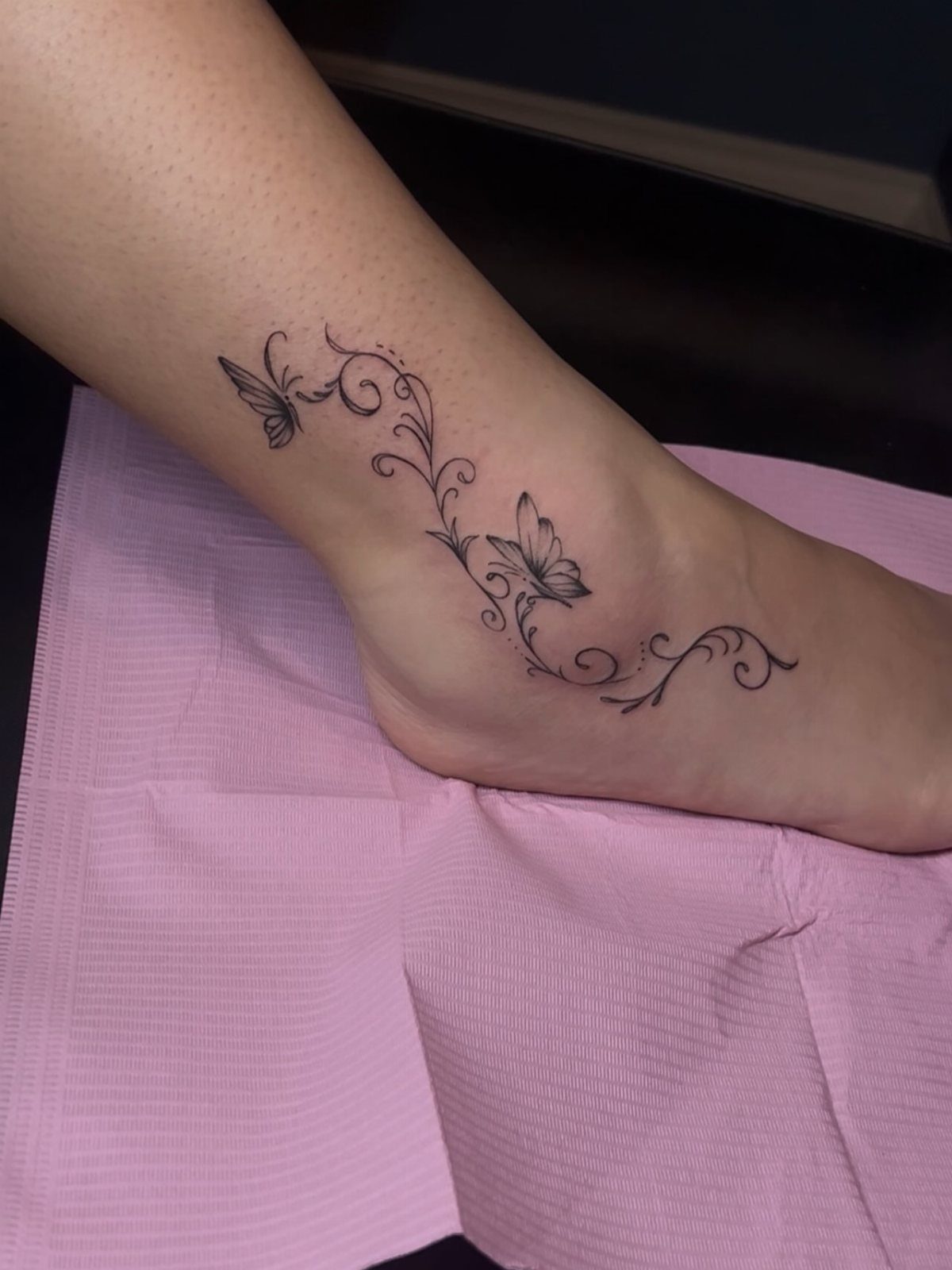 A delicate black ink butterfly and swirl tattoo on a person's ankle and foot.