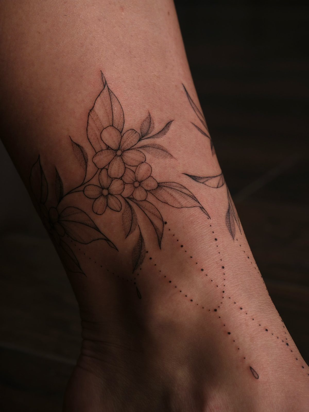 A floral tattoo with delicate leaves and dotted lines wraps around a person's ankle.
