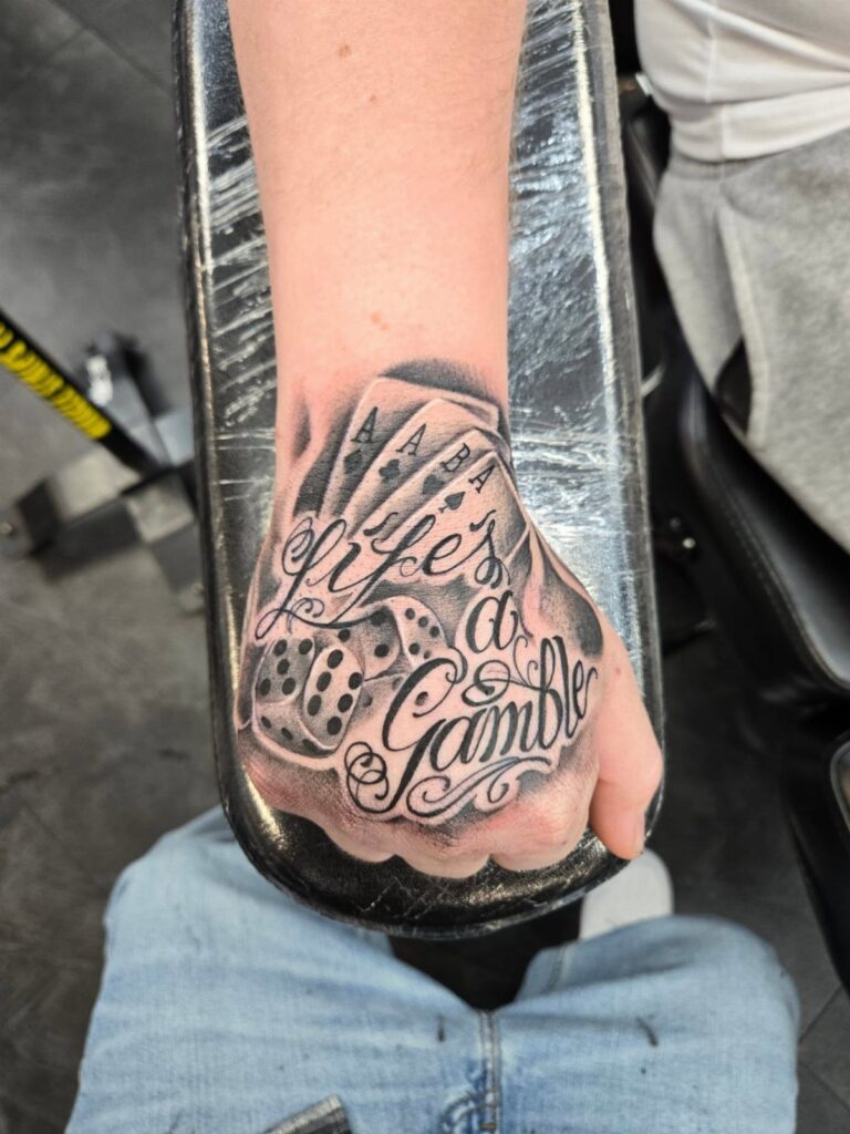 A person's hand displaying a large black and grey tattoo of playing cards, dice, and script text.