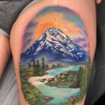 A colorful mountain landscape tattoo on skin featuring a snowy peak, river, and pine trees.