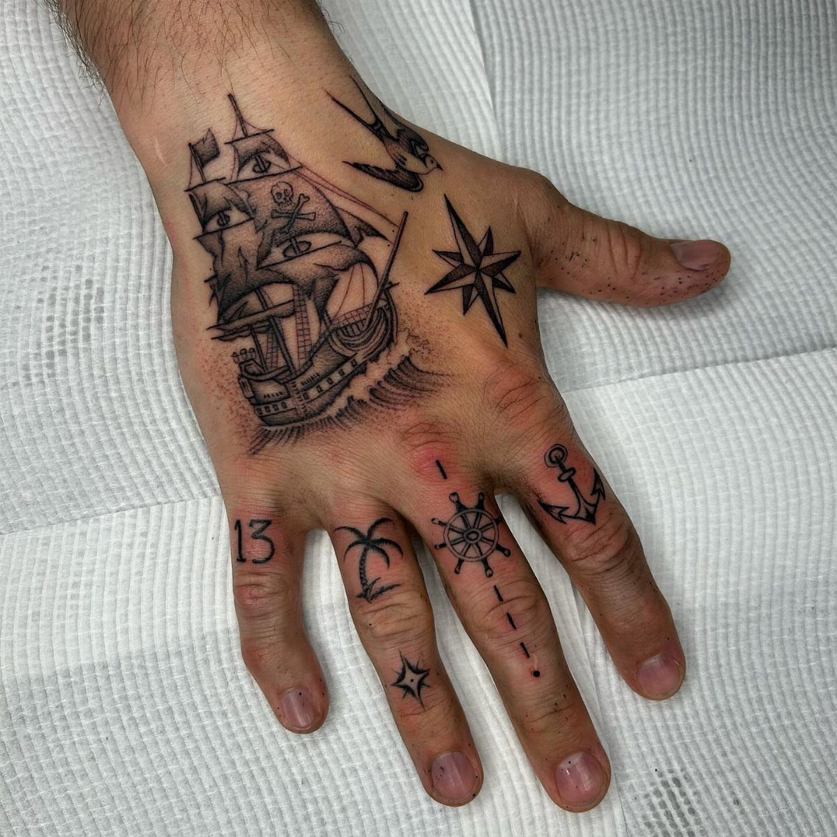 A person's hand decorated with various black ink nautical tattoos including a ship and stars.