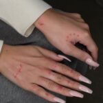 Hands with long pink nails featuring several small, delicate red fine line tattoos on the fingers and knuckles.