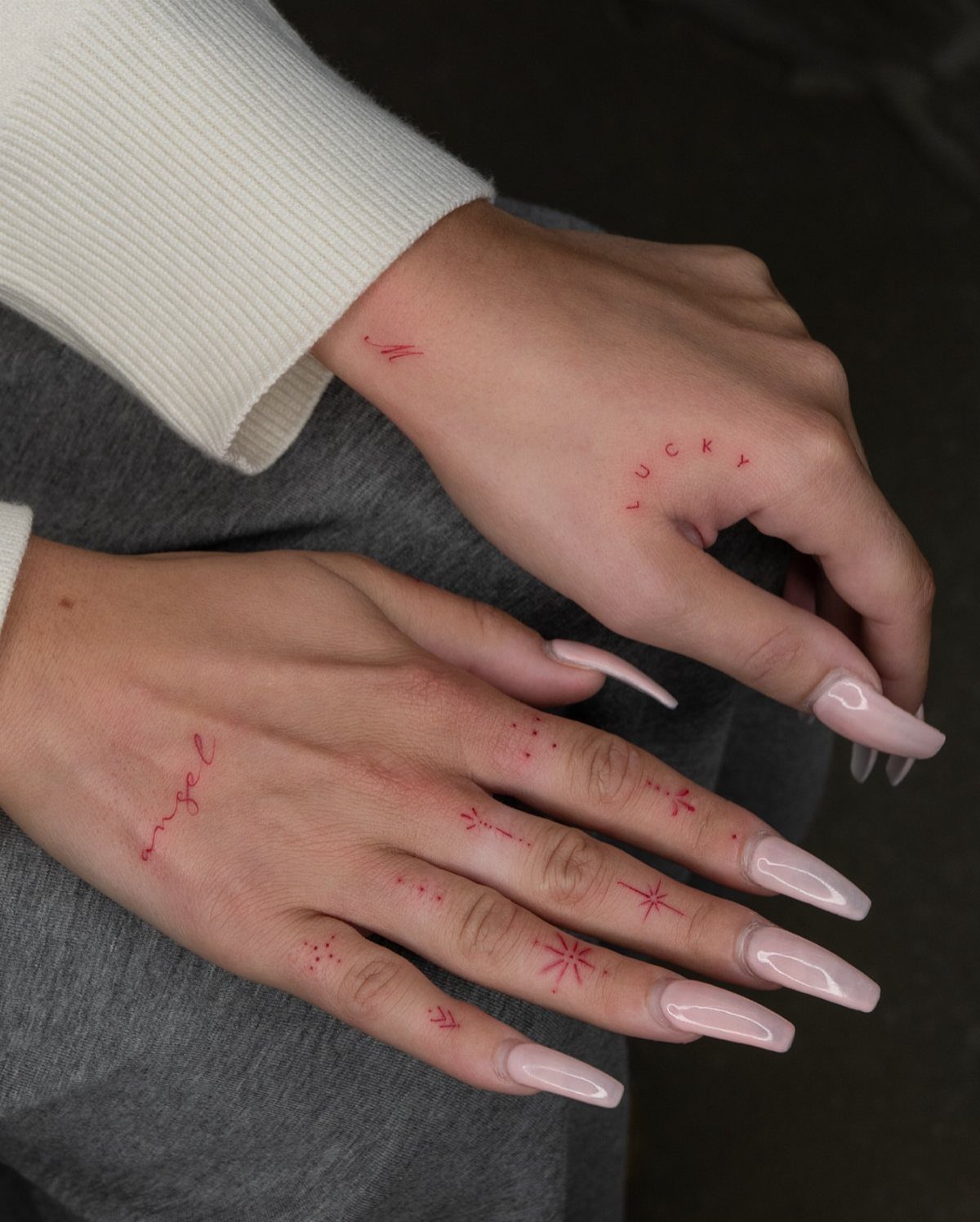 Hands with long pink nails featuring several small, delicate red fine line tattoos on the fingers and knuckles.