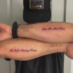 Two male arms crossed, displaying small script tattoos reading 'This Too Shall Pass' and 'In God's Waiting Room'.