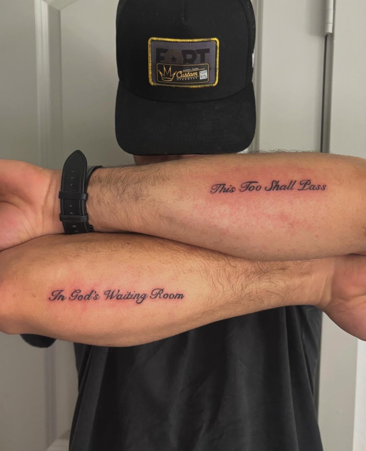 Two male arms crossed, displaying small script tattoos reading 'This Too Shall Pass' and 'In God's Waiting Room'.