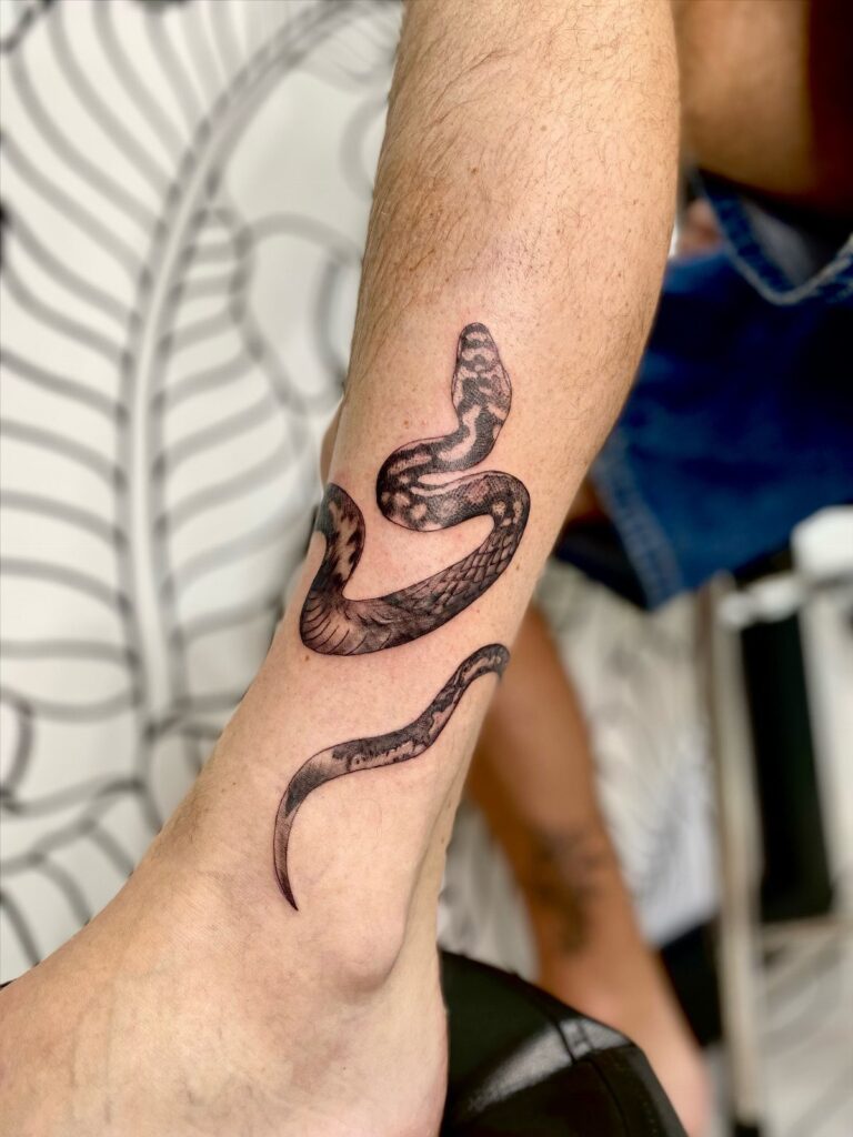 A black ink snake tattoo wrapped around a person's lower leg and ankle.