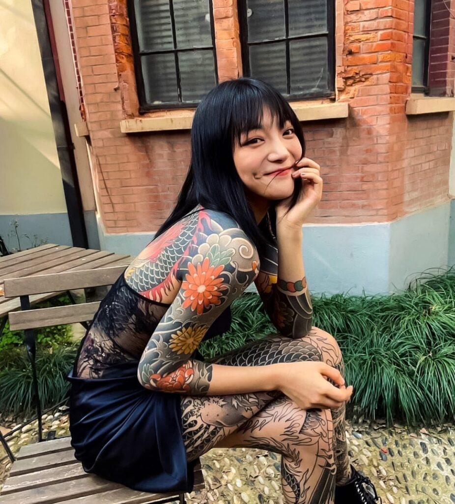 A woman sitting outdoors displaying large, colorful Japanese-style tattoos on her arm and leg.