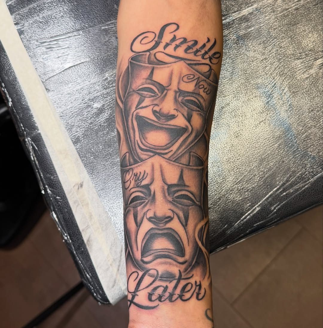 Black and grey realism tattoo of comedy and tragedy masks on a person's forearm.