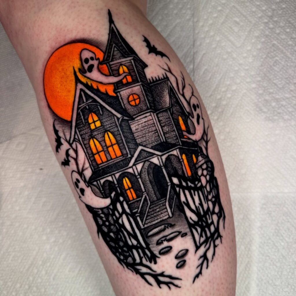 A spooky haunted house tattoo with orange glowing windows and ghosts on a person's leg.