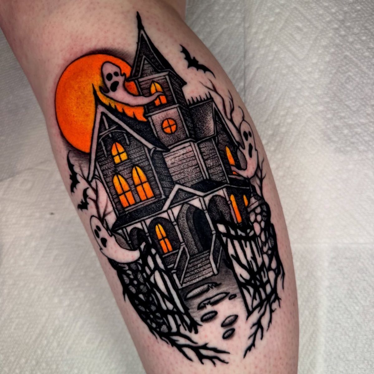 A spooky haunted house tattoo with orange glowing windows and ghosts on a person's leg.