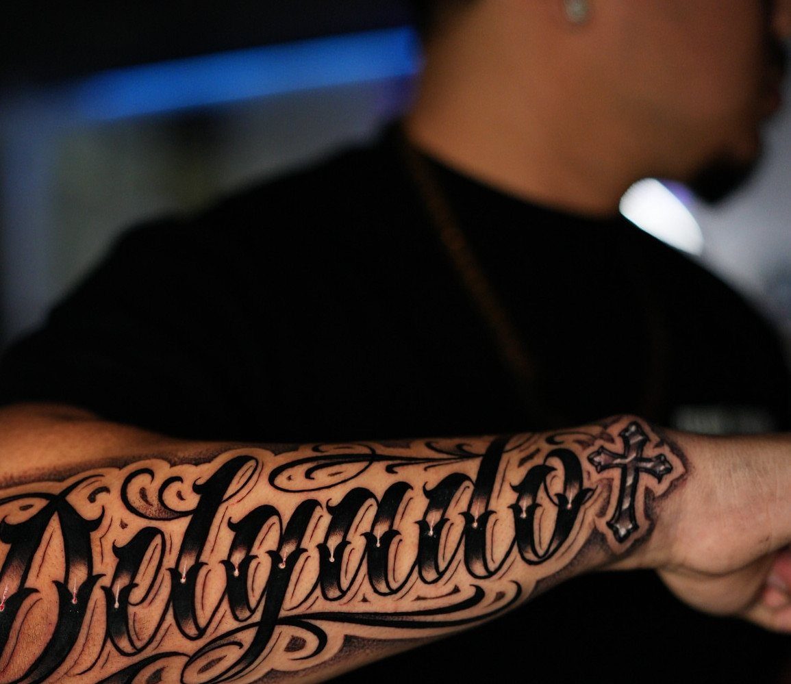 A detailed black and grey tattoo of stylized script lettering and a cross on a person's forearm.