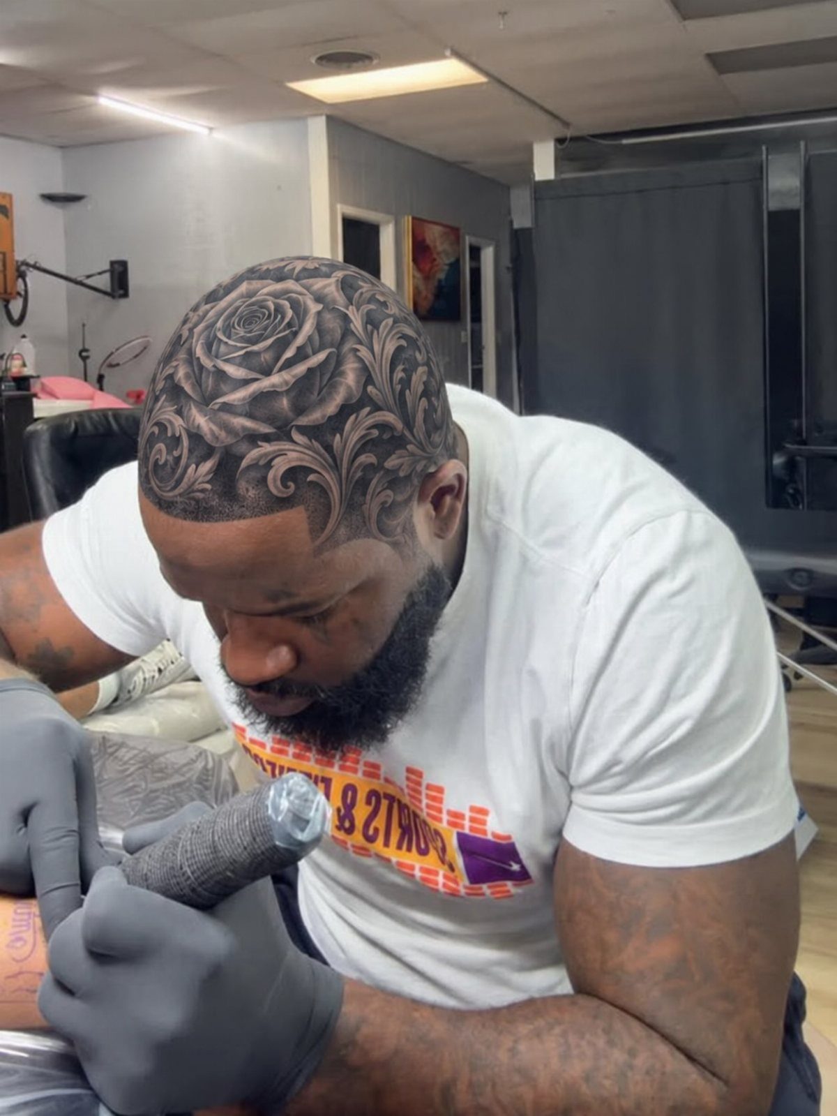 A man tattooing an intricate rose and acanthus leaf pattern onto a client's shaved head.